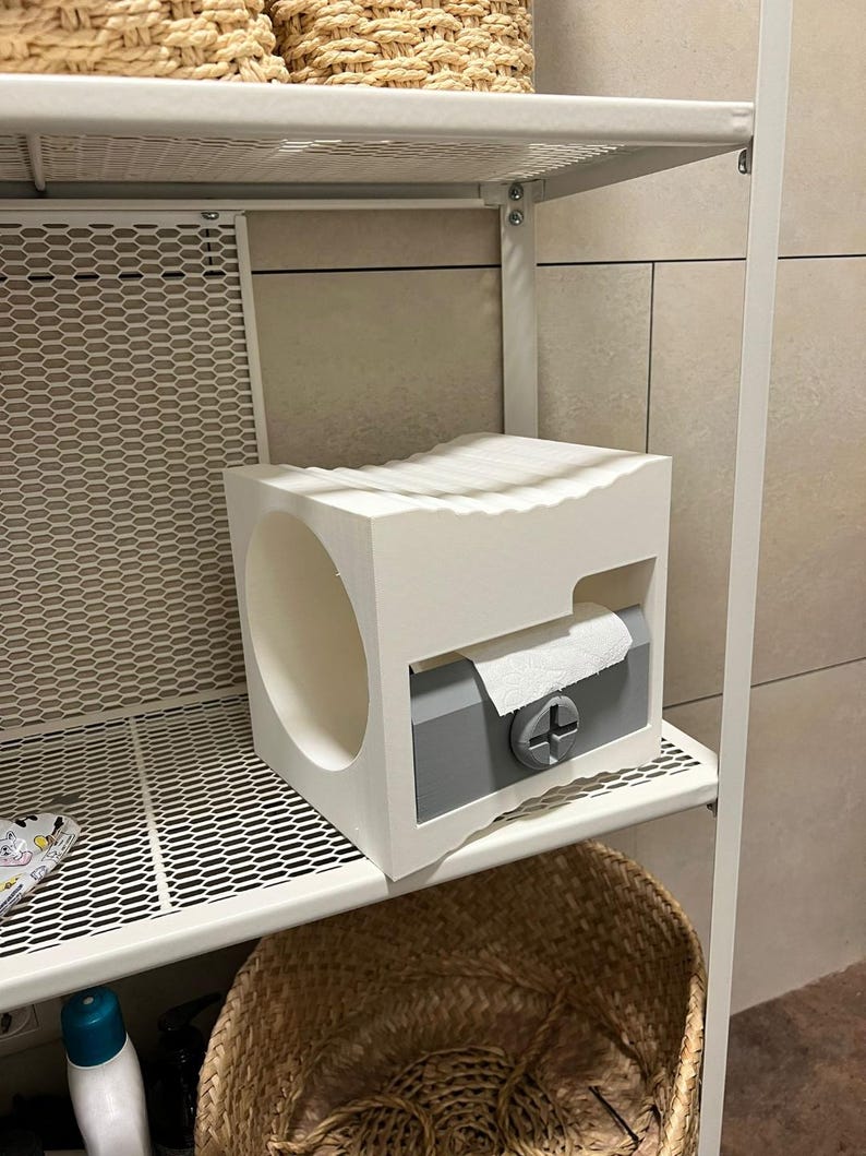 May include: A white and gray toilet paper holder with a circular opening and a slot for dispensing paper. The holder is on a white metal shelf, with a woven basket below. The design is modern and minimalist.