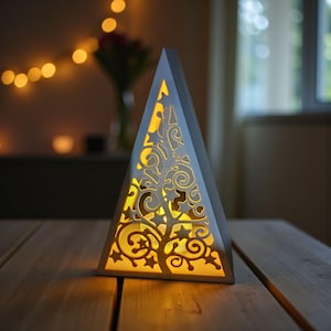 May include: A white, pyramid-shaped decorative lantern with a tree and star design cut into the sides. The interior glows with a warm yellow light, creating a cozy ambiance. The lantern sits on a wooden table.