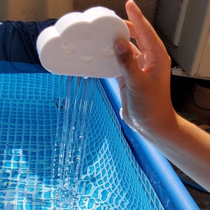 May include: A white cloud-shaped object with a smiling face is held over a blue pool, creating a stream of water. The pool has a blue and white checkered pattern. The image is taken outdoors in daylight.