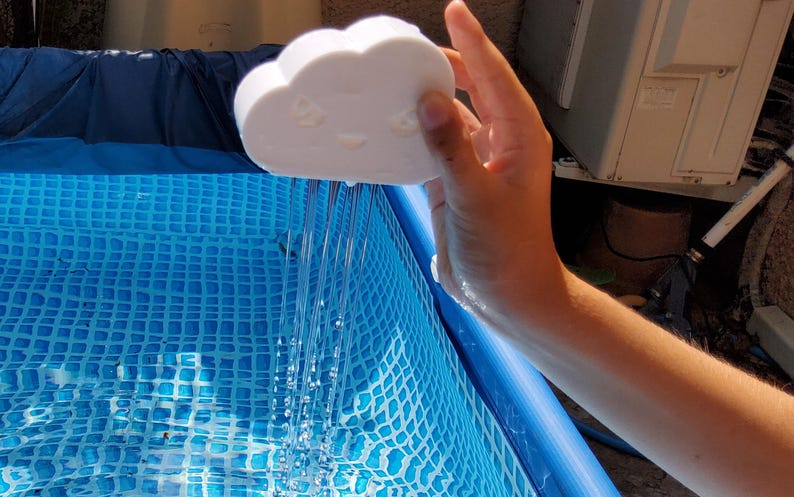 May include: A white cloud-shaped object with a smiling face is held over a blue pool, creating a stream of water. The pool has a blue and white checkered pattern. The image is taken outdoors in daylight.
