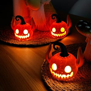 May include: Three glowing orange Halloween pumpkin decorations with devil horns and a carved, toothy grin. Each pumpkin sits on a woven mat, with a black curly stem. The pumpkins are lit from within, creating a spooky ambiance.