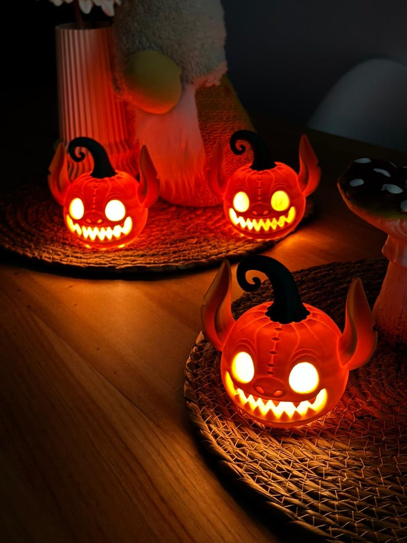 May include: Three glowing orange Halloween pumpkin decorations with devil horns and a carved, toothy grin. Each pumpkin sits on a woven mat, with a black curly stem. The pumpkins are lit from within, creating a spooky ambiance.