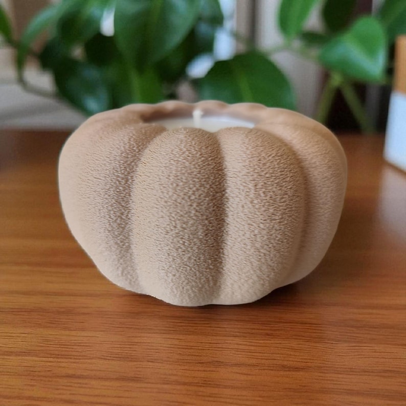 May include: A tan pumpkin-shaped candle with a textured surface. The candle has a white wick and sits on a wooden surface. The background includes green foliage.