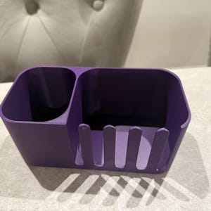 May include: A solid purple desk organizer with two compartments and a slotted front. The organizer is made of a smooth material and sits on a light-colored surface. The design is simple and functional.