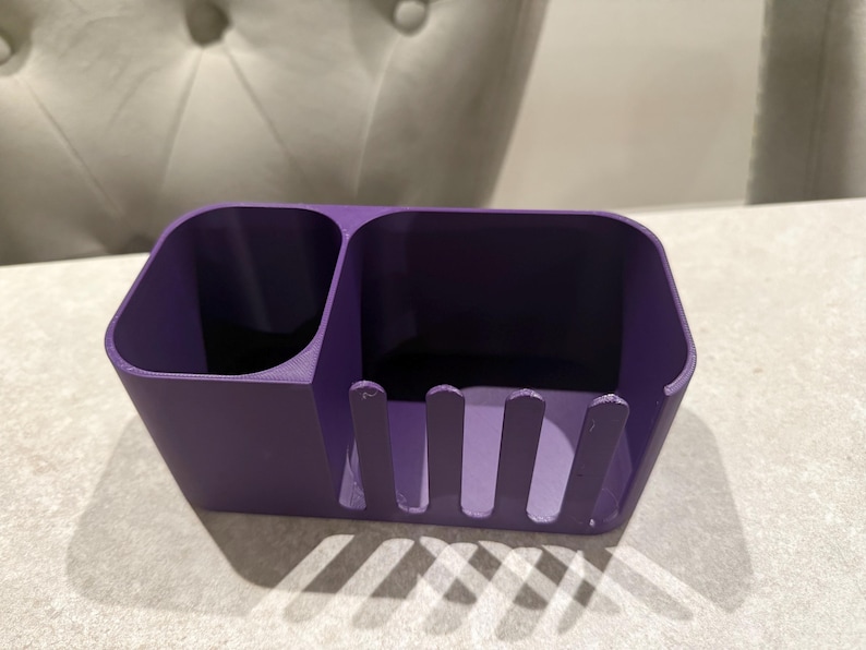 May include: A solid purple desk organizer with two compartments and a slotted front. The organizer is made of a smooth material and sits on a light-colored surface. The design is simple and functional.