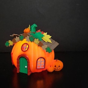 K&ouml;nnte beinhalten: Ein 3D-gedrucktes orangefarbenes K&uuml;rbis-Haus mit einer gr&uuml;nen T&uuml;r, Fenstern und einem Dach, das mit bunten Bl&auml;ttern verziert ist. Zwei kleine K&uuml;rbisse stehen daneben. Das Haus hat einen warmen Schein, ideal f&uuml;r Halloween-Dekorationen.