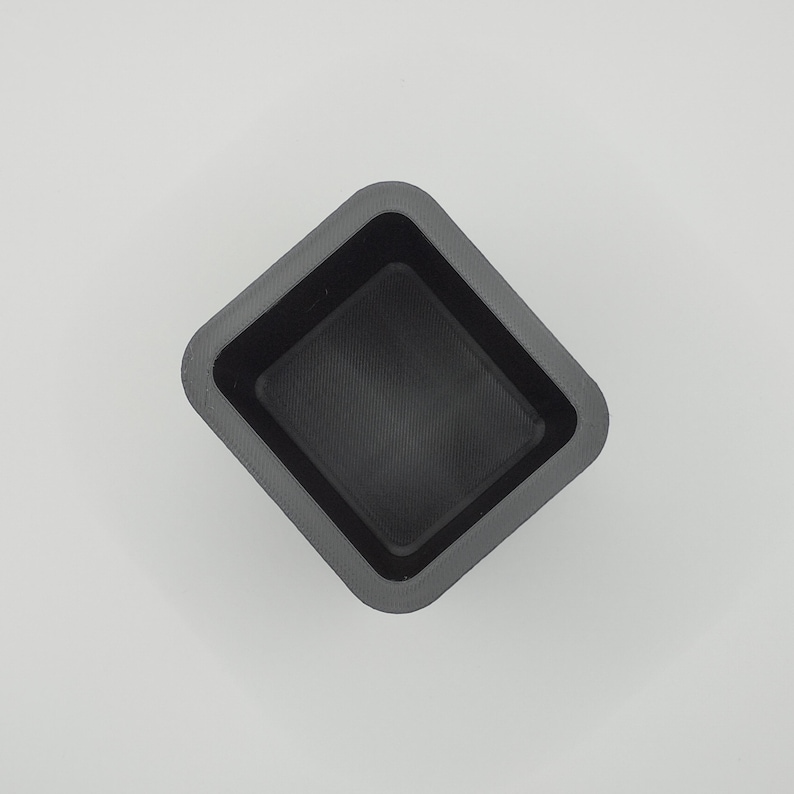 May include: A dark gray, square-shaped container with a slightly textured interior. The container has a raised, angled rim and a flat bottom. The object is set against a plain white background.