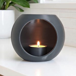 May include: A dark grey, spherical candle holder with a large circular aperture. A lit tealight candle sits within, casting a warm glow. The holder is made of a matte material and is placed on a white surface.