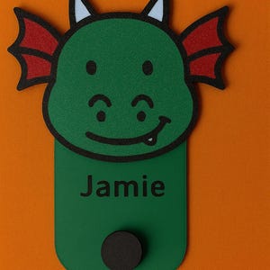 May include: A green dragon-shaped name tag with the name "Jamie" printed in black. The dragon has red wings, blue accents on its horns, and a smiling face. A black circular magnet is at the bottom.