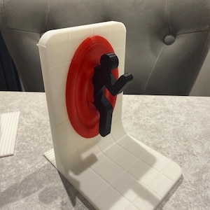 May include: A white, 3D-printed desk organizer featuring a red circular portal and a black silhouette of a person. The organizer has a rectangular base and a vertical back with a grid pattern.