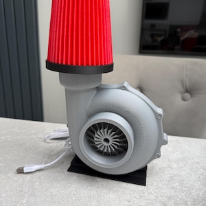 May include: A unique desk lamp featuring a gray turbine housing with a red air filter shade. The lamp has a USB cable for power and sits on a black base. The design is reminiscent of automotive parts.