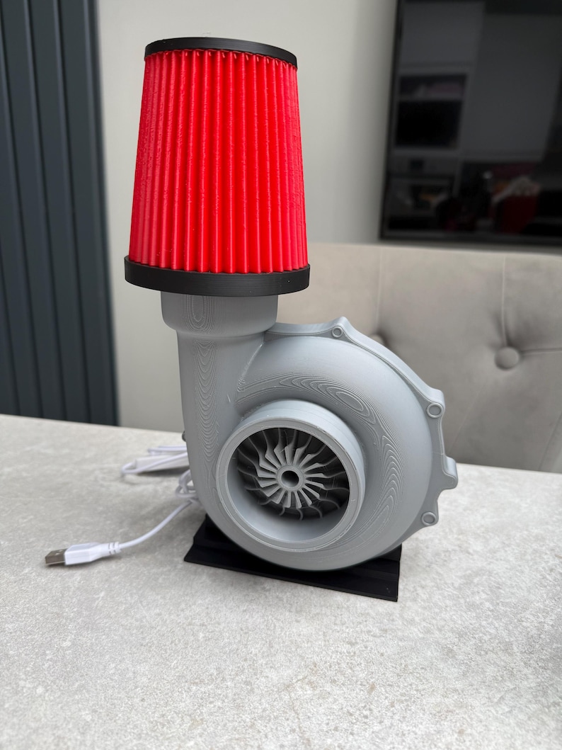 May include: A unique desk lamp featuring a gray turbine housing with a red air filter shade. The lamp has a USB cable for power and sits on a black base. The design is reminiscent of automotive parts.
