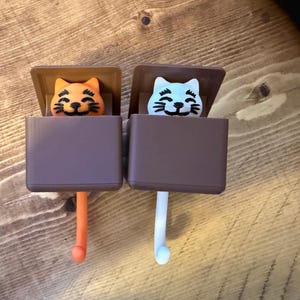 May include: Two cat-shaped wall hooks. One hook features an orange cat, the other a light blue cat. Both cats have black whiskers and are set against a brown background. The hooks are brown and have a rounded end.