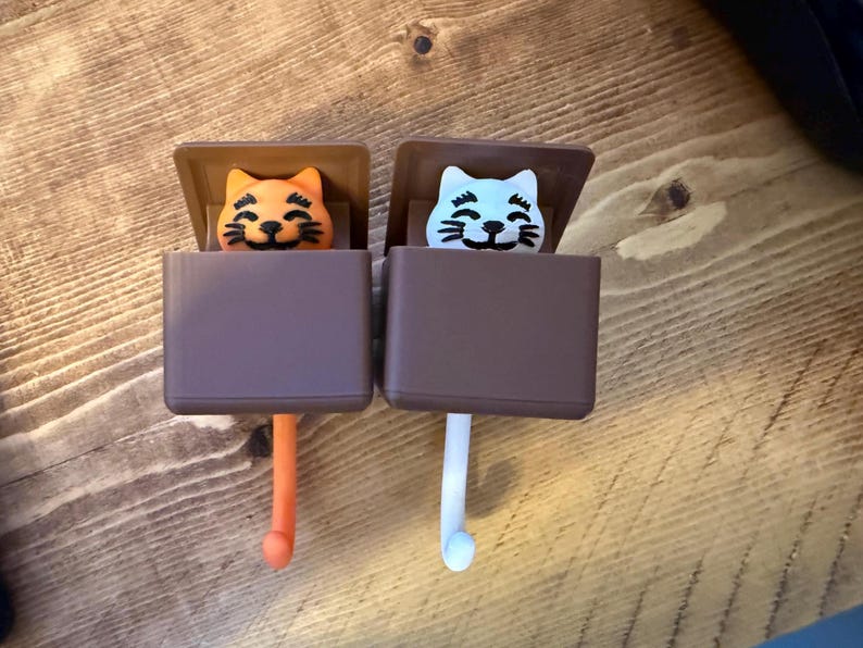 May include: Two cat-shaped wall hooks. One hook features an orange cat, the other a light blue cat. Both cats have black whiskers and are set against a brown background. The hooks are brown and have a rounded end.