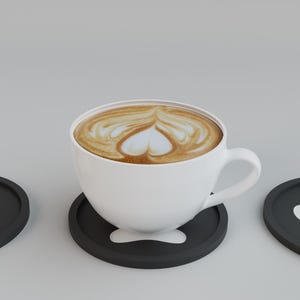 May include: A white ceramic coffee cup with latte art in the shape of a heart, resting on a black coaster. Two additional black coasters with white paw print designs are on either side. The coffee has a creamy, light brown color.