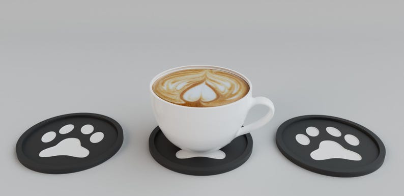 May include: A white ceramic coffee cup with latte art in the shape of a heart, resting on a black coaster. Two additional black coasters with white paw print designs are on either side. The coffee has a creamy, light brown color.