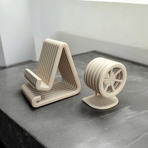 Puede incluir: Dos soportes de tel&eacute;fono de madera. Uno tiene un dise&ntilde;o en forma de A y el otro tiene forma de rueda. Ambos son de madera clara y est&aacute;n sobre una superficie gris. El soporte en forma de A mide unos 15 cm de alto.