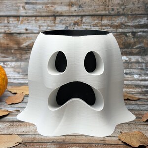 May include: A white ghost-shaped planter with a black interior, featuring large eye and mouth cutouts. The planter is on a wooden surface with a pumpkin and autumn leaves, perfect for Halloween decor.