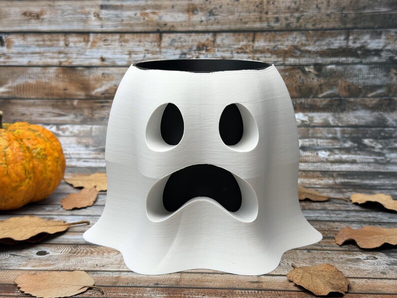 May include: A white ghost-shaped planter with a black interior, featuring large eye and mouth cutouts. The planter is on a wooden surface with a pumpkin and autumn leaves, perfect for Halloween decor.