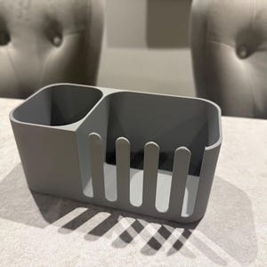 May include: A gray plastic utensil holder with two compartments. One compartment is a small, round cup, and the other has vertical slots. The holder is on a light-colored surface, with shadows cast by the object.