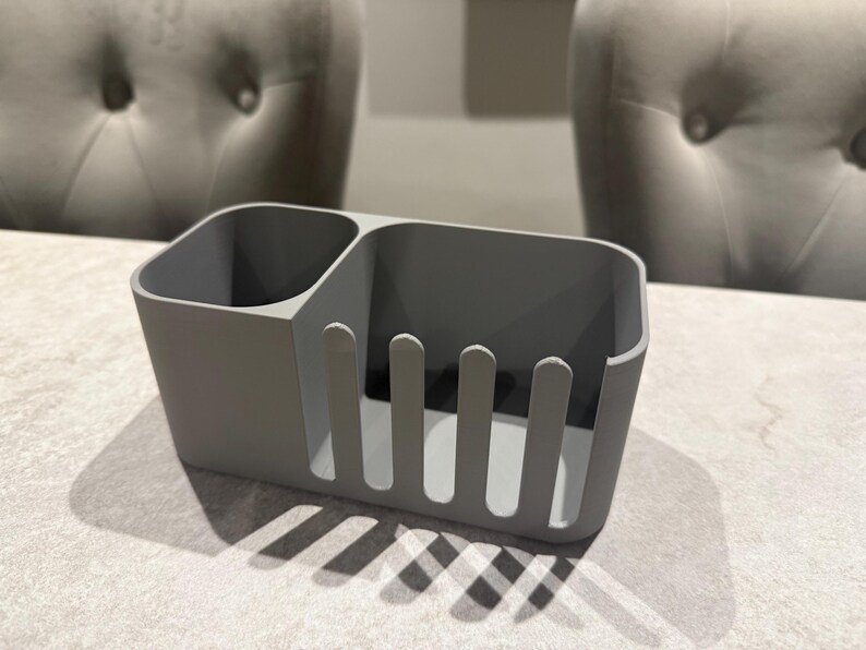 May include: A gray plastic utensil holder with two compartments. One compartment is a small, round cup, and the other has vertical slots. The holder is on a light-colored surface, with shadows cast by the object.