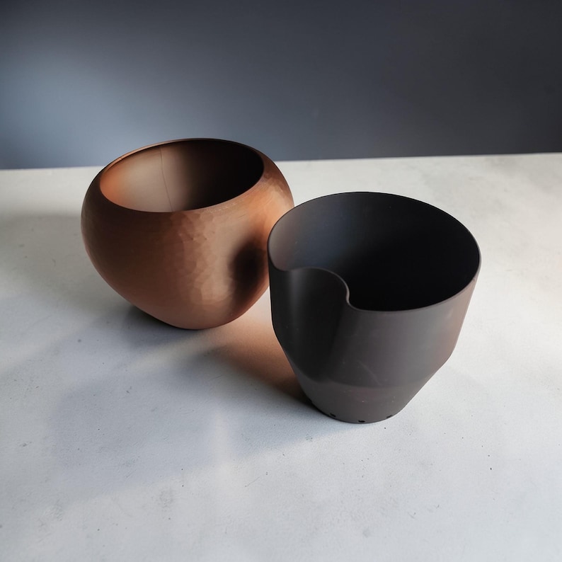 May include: Two decorative bowls. One is a hammered copper color, spherical, and open at the top. The other is a dark brown, asymmetrical shape with a curved opening. Both sit on a light-colored surface.