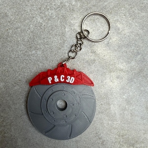 May include: A gray and red 3D-printed keychain featuring a brake rotor design. The red section at the top has the text "P & C 3D" in white. The keychain includes a silver chain and a keyring.