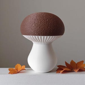 May include: A ceramic mushroom-shaped vase with a textured brown top and a white stem. The vase has a ribbed design at the base of the cap. Two orange leaves are placed near the vase.