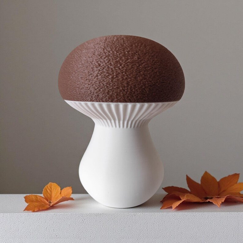 May include: A ceramic mushroom-shaped vase with a textured brown top and a white stem. The vase has a ribbed design at the base of the cap. Two orange leaves are placed near the vase.