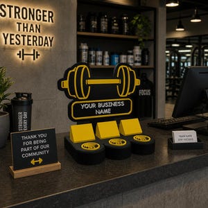 May include: A gym counter display featuring a black and yellow dumbbell logo with the text "YOUR BUSINESS NAME." Additional signs read "STRONGER THAN YESTERDAY" and "THANK YOU FOR BEING PART OF OUR COMMUNITY."