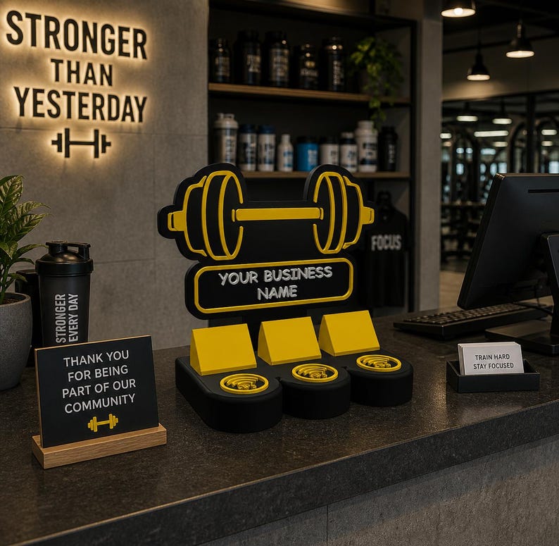 May include: A gym counter display featuring a black and yellow dumbbell logo with the text "YOUR BUSINESS NAME." Additional signs read "STRONGER THAN YESTERDAY" and "THANK YOU FOR BEING PART OF OUR COMMUNITY."