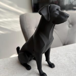 May include: A black Labrador Retriever statue. The dog is sitting with its head turned to the right. The statue has a smooth, matte finish and is placed on a light-colored surface.