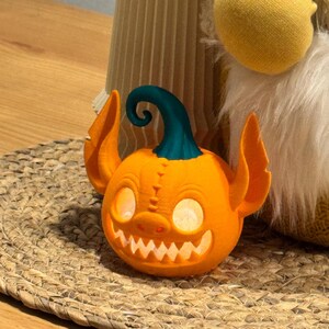 May include: A 3D-printed orange pumpkin with a teal stem and large, pointed ears. The pumpkin has a carved face with glowing eyes and a toothy grin. It is sitting on a woven placemat.