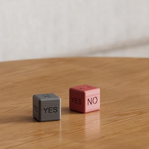 May include: Two small, cube-shaped decision makers sit on a wooden surface. One is gray and says "YES" on two sides. The other is red and says "YES" and "NO" on adjacent sides. The cubes are for making decisions.
