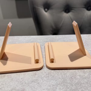 May include: Two peach-colored, angled phone stands. Each stand has a rectangular base with a rounded corner and a vertical support. The support is a thin, rectangular piece with a lighter-colored, angled top. The stands are on a gray surface.