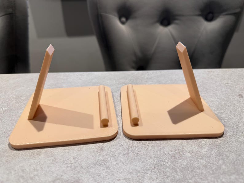 May include: Two peach-colored, angled phone stands. Each stand has a rectangular base with a rounded corner and a vertical support. The support is a thin, rectangular piece with a lighter-colored, angled top. The stands are on a gray surface.