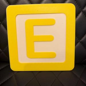May include: A yellow square block with a white center and a yellow letter "E". The letter is centered within the white space. The block has rounded corners.