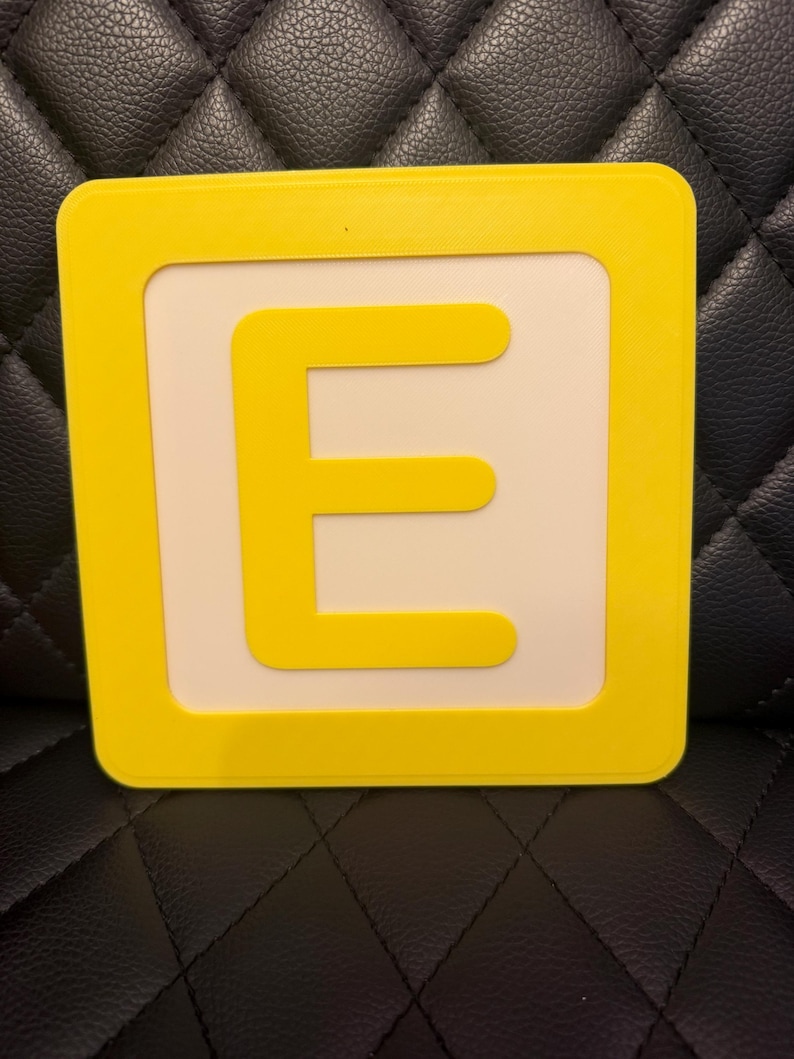 May include: A yellow square block with a white center and a yellow letter "E". The letter is centered within the white space. The block has rounded corners.