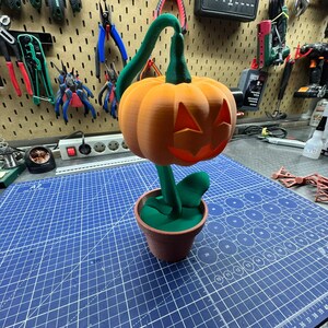 May include: A 3D-printed Halloween decoration featuring an orange jack-o'-lantern with carved eyes and a mouth. The pumpkin sits atop a green stem and leaves, planted in a brown pot. The background shows tools and a blue cutting mat.
