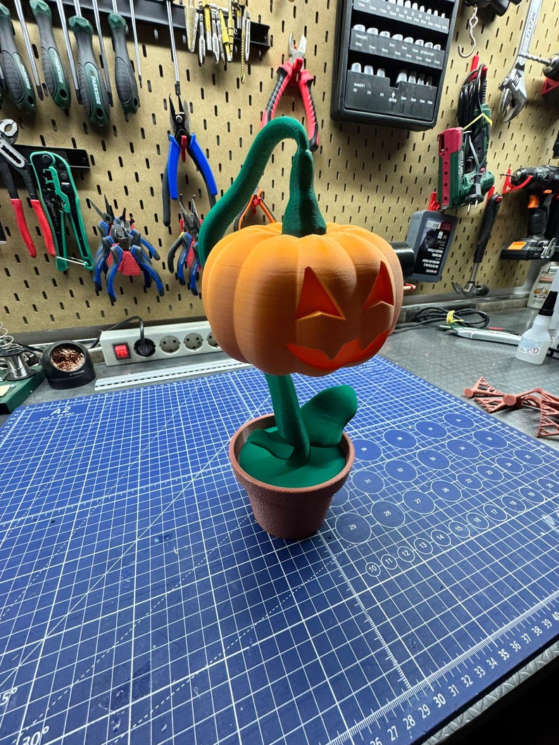 May include: A 3D-printed Halloween decoration featuring an orange jack-o'-lantern with carved eyes and a mouth. The pumpkin sits atop a green stem and leaves, planted in a brown pot. The background shows tools and a blue cutting mat.