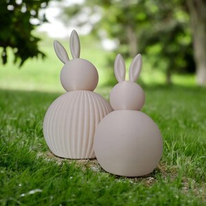 May include: Two light pink bunny sculptures on green grass. One bunny has a ribbed body, and the other has a smooth body. Both have long ears and a round head. The background is blurred with trees.