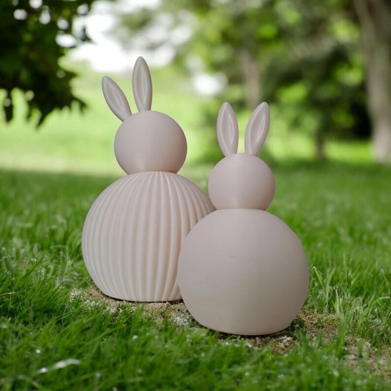 May include: Two light pink bunny sculptures on green grass. One bunny has a ribbed body, and the other has a smooth body. Both have long ears and a round head. The background is blurred with trees.