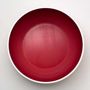 May include: A top-down view of a round, red bowl with a white rim. The bowl's interior is a solid, vibrant red, and the white rim provides a clean contrast. The bowl is likely made of ceramic or a similar material.