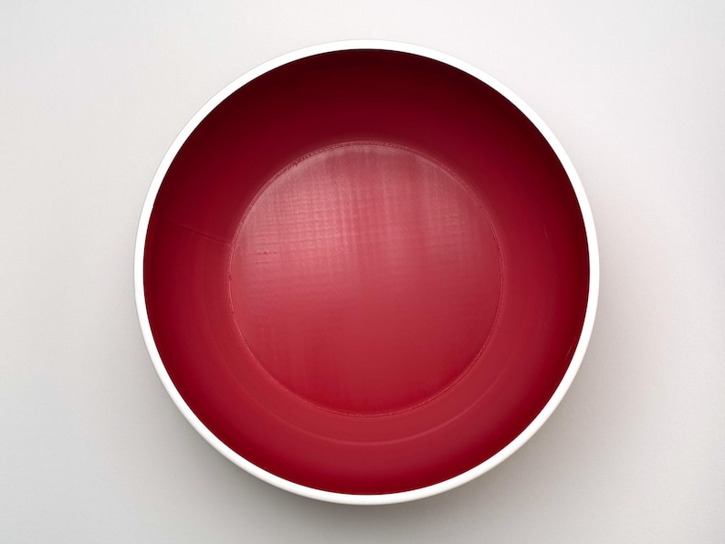 May include: A top-down view of a round, red bowl with a white rim. The bowl's interior is a solid, vibrant red, and the white rim provides a clean contrast. The bowl is likely made of ceramic or a similar material.