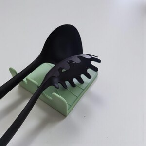 Op de afbeelding: Een zwarte siliconen lepel en pastaserver rusten op een lichtgroene keukengerei houder. De pastaserver heeft een uniek, golvend ontwerp. Keukengerei voor koken en serveren.