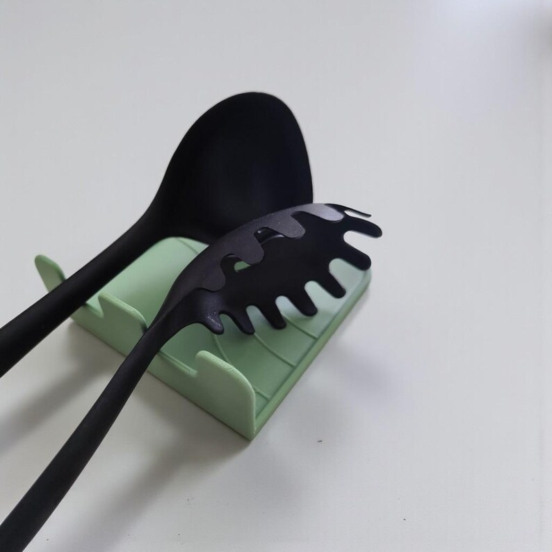 Op de afbeelding: Een zwarte siliconen lepel en pastaserver rusten op een lichtgroene keukengerei houder. De pastaserver heeft een uniek, golvend ontwerp. Keukengerei voor koken en serveren.
