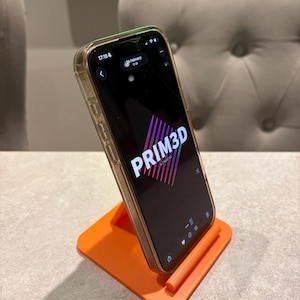 May include: An orange 3D-printed phone stand holding a smartphone. The phone displays the text "PRIM3D" in pink and purple. The phone has a clear case. The stand is on a light gray surface.