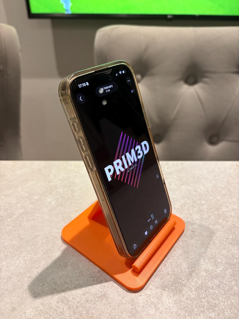 May include: An orange 3D-printed phone stand holding a smartphone. The phone displays the text "PRIM3D" in pink and purple. The phone has a clear case. The stand is on a light gray surface.