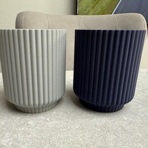 May include: Two cylindrical planters with vertical ribbed detailing. One is light gray, the other is navy blue. Both have a slightly flared base. The planters are on a light gray surface, suggesting a modern home decor theme.