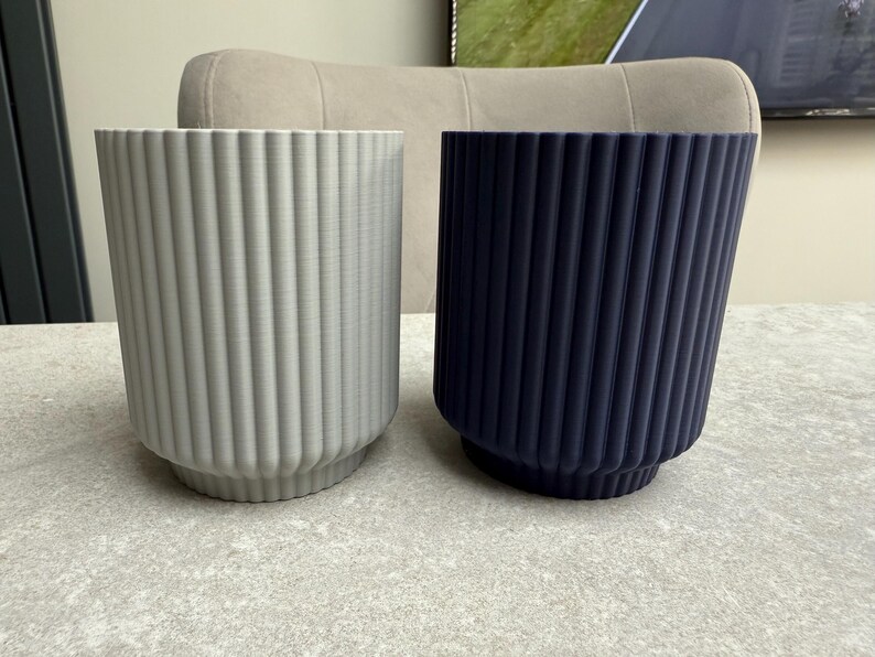 May include: Two cylindrical planters with vertical ribbed detailing. One is light gray, the other is navy blue. Both have a slightly flared base. The planters are on a light gray surface, suggesting a modern home decor theme.